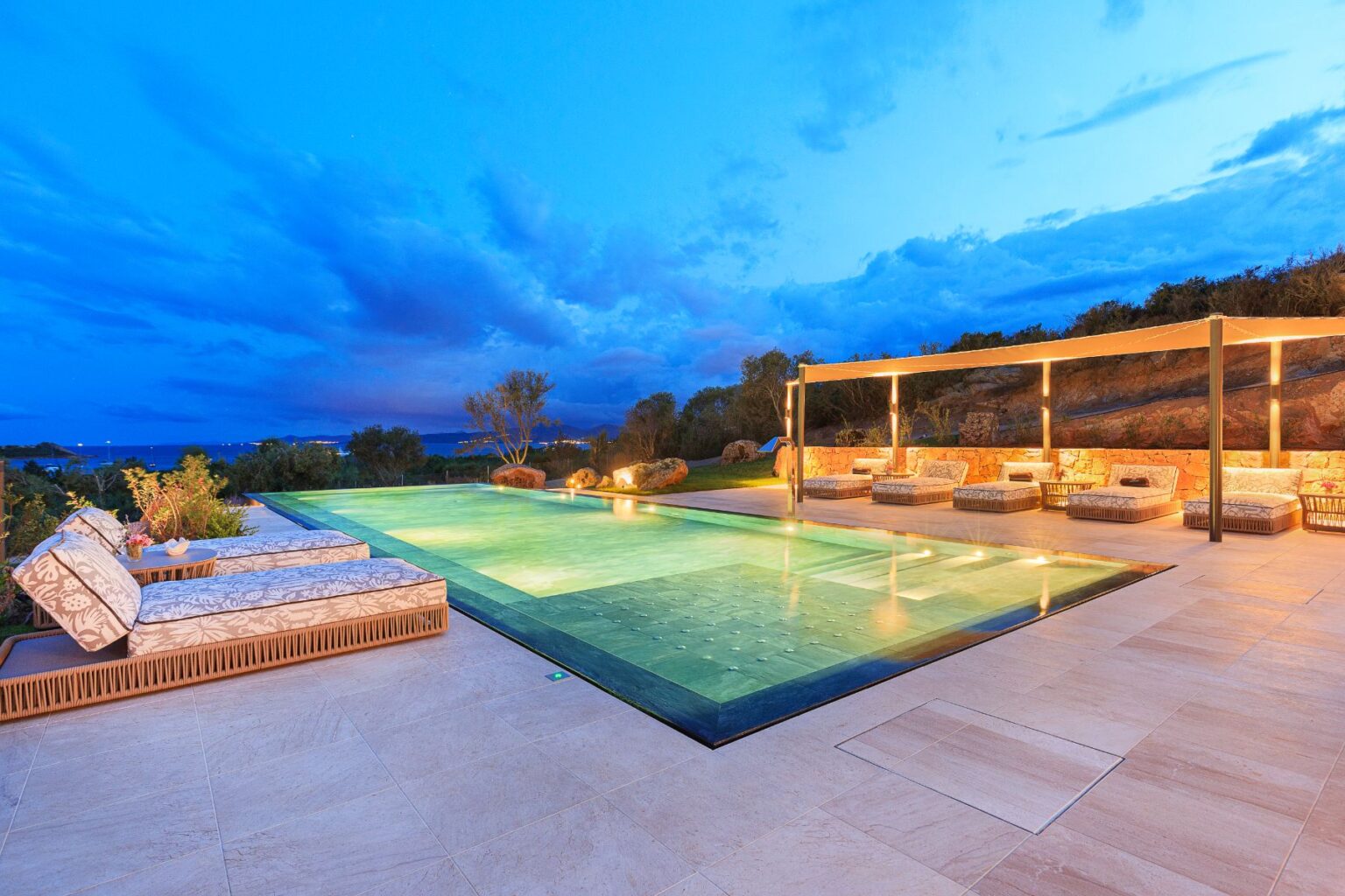 Luxurious outdoor swimming pool surrounded by stylish lounge chairs, illuminated by soft lighting at dusk, with a scenic view of the ocean and lush greenery in the background. | Only on SopranoVillas