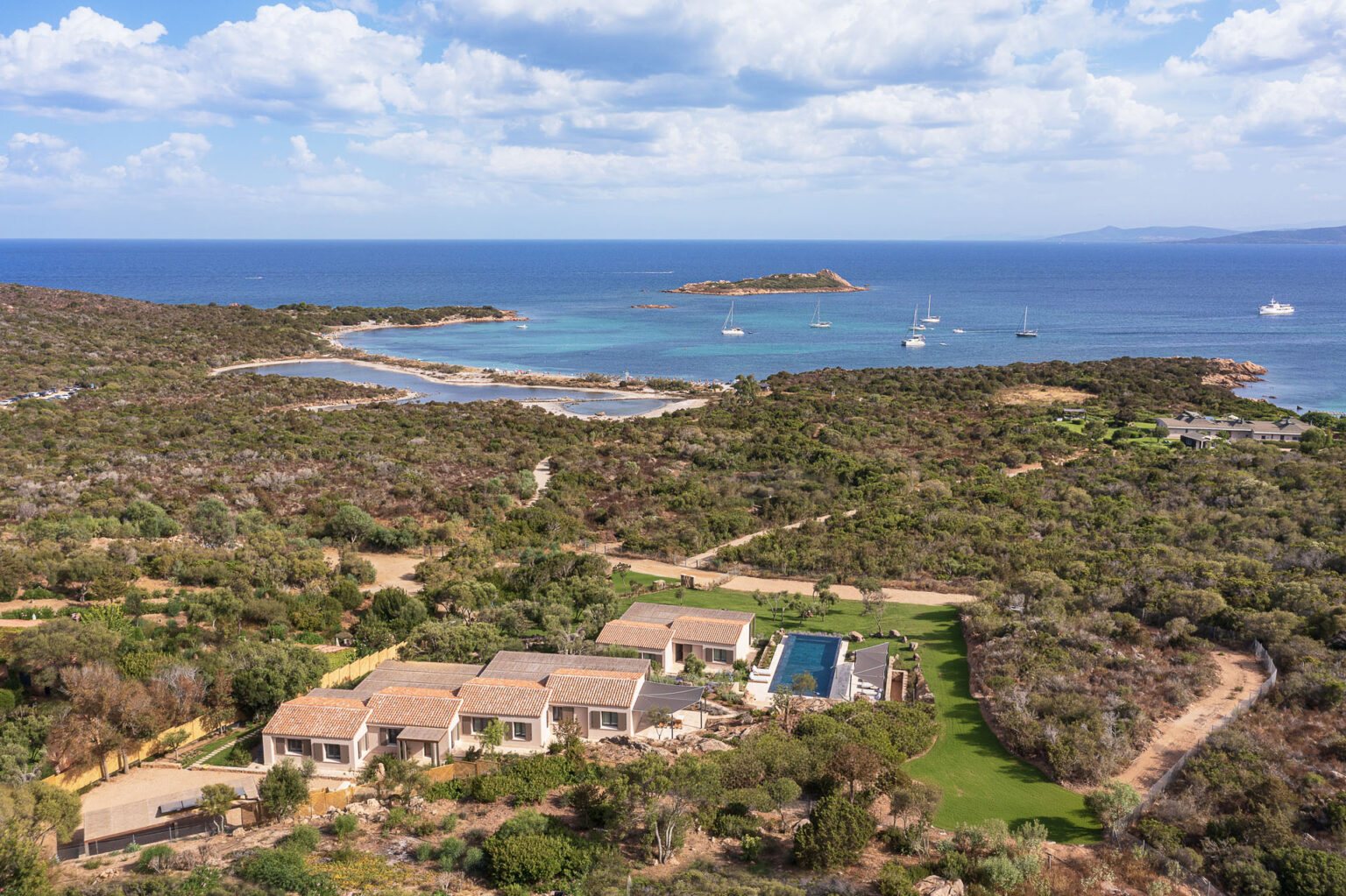Aerial view of a coastal landscape featuring a luxurious villa with a swimming pool surrounded by lush greenery, overlooking a serene blue sea dotted with sailboats and a small island in the distance. Ideal for vacation and relaxation. | Only on SopranoVillas