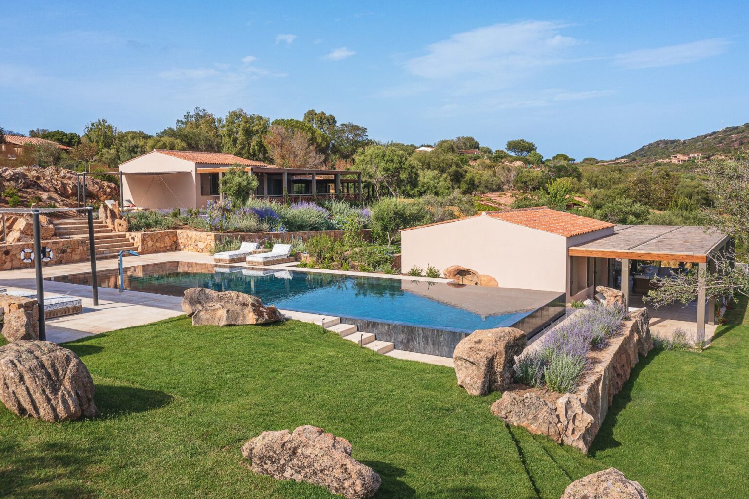Stunning modern villa with a sleek infinity pool, surrounded by lush greenery and natural stone features, set against a clear blue sky. The outdoor space includes comfortable lounge chairs and beautifully landscaped gardens, ideal for relaxation and luxury living. | Only on SopranoVillas