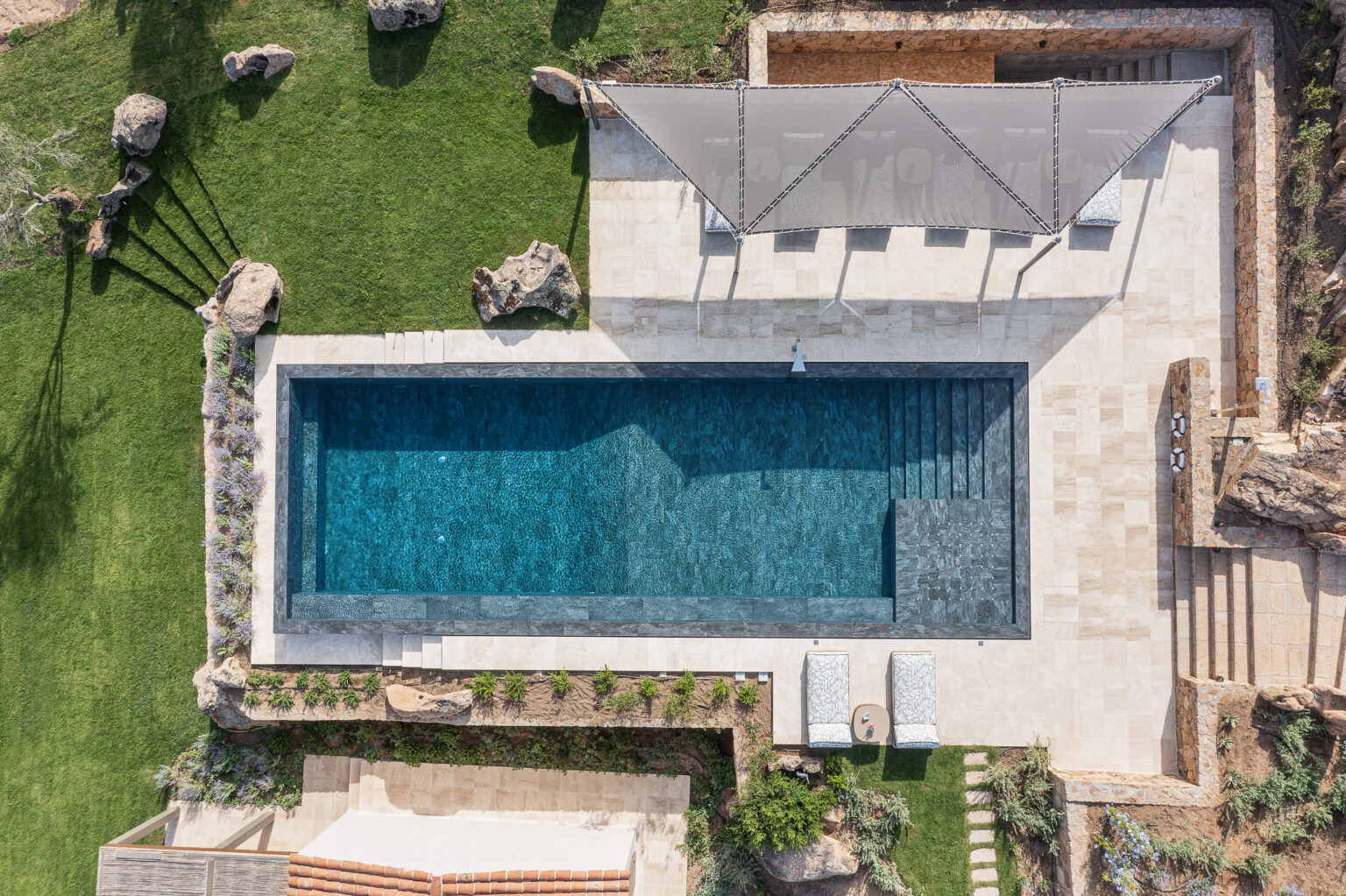 Aerial view of a modern swimming pool surrounded by lush green grass, stone landscaping, and a shaded seating area with lounge chairs. The pool features a sleek design with dark tiles and steps leading into the water, complemented by natural rock formations and ornamental plants. Perfect for outdoor relaxation and summer enjoyment. | Only on SopranoVillas