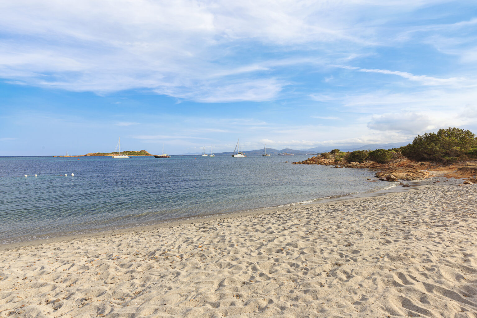 Scenic beach view featuring soft sandy shores, calm blue waters, and anchored sailboats in the distance under a bright sky with scattered clouds. | Only on SopranoVillas