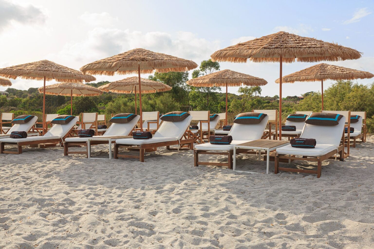 Beachside lounge chairs with straw umbrellas, surrounded by soft sand and greenery, create a relaxing atmosphere for sunbathing and leisure. | Only on SopranoVillas