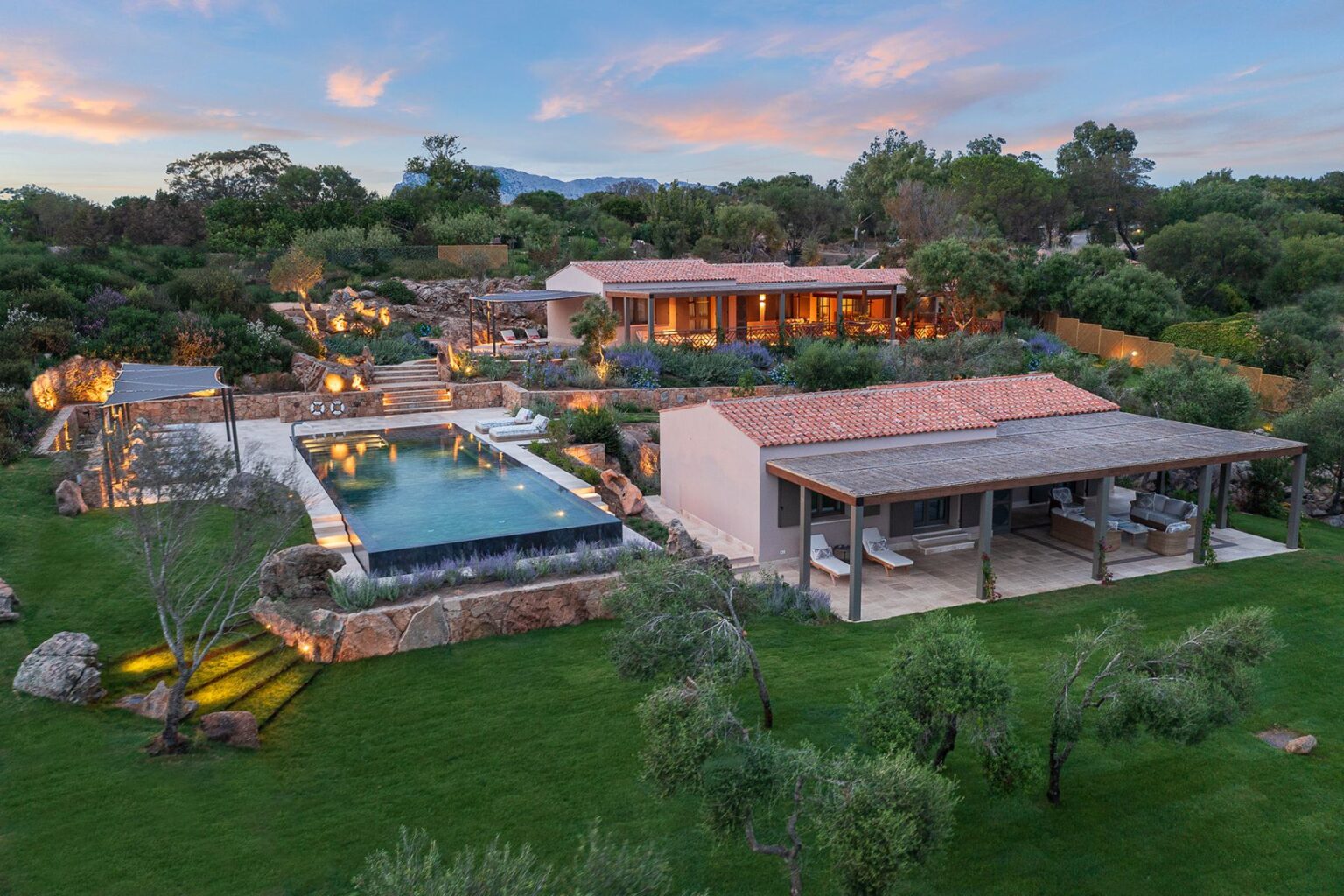 Aerial view of a luxurious villa featuring a spacious outdoor area with a modern infinity pool, landscaped gardens, and multiple seating areas. The property is surrounded by lush greenery and rocky terrain, showcasing a blend of natural beauty and elegant architecture at sunset. | Only on SopranoVillas