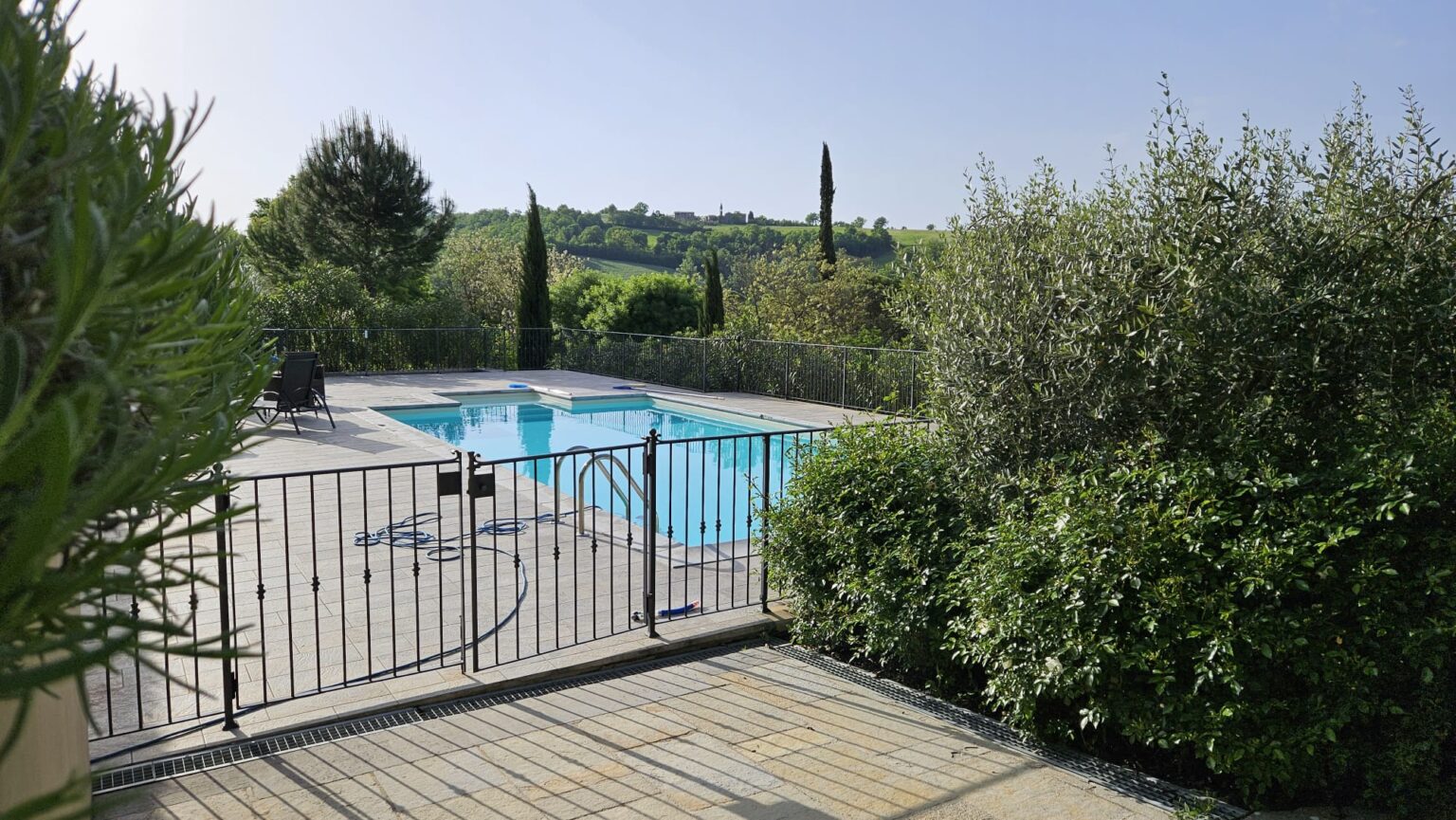Scenic view of a private swimming pool surrounded by lush greenery and a black metal fence, with rolling hills in the background under a clear blue sky. | Only on SopranoVillas