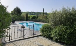Scenic view of a private swimming pool surrounded by lush greenery and a black metal fence, with rolling hills in the background under a clear blue sky.