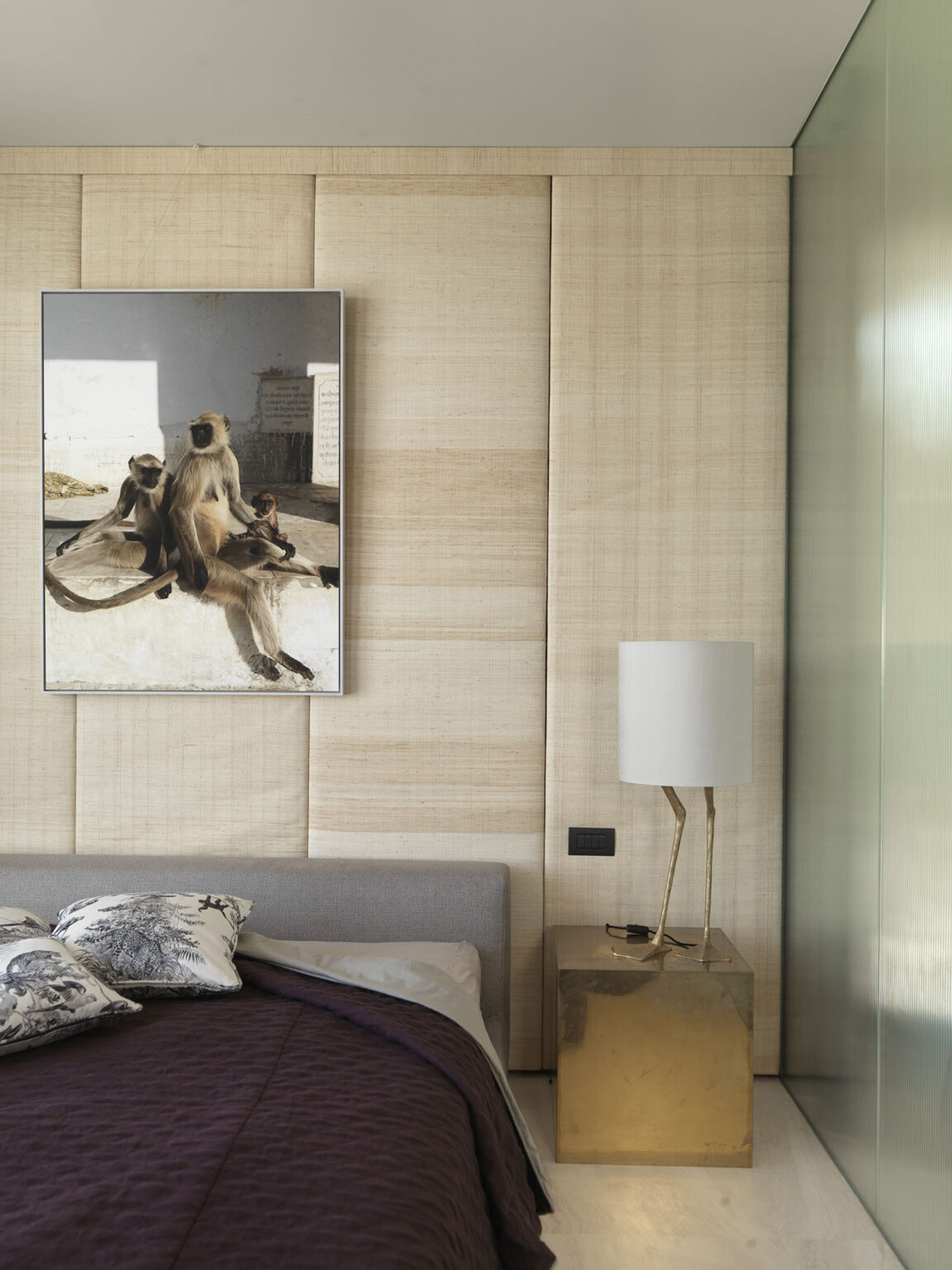 Modern bedroom interior featuring a gray bed with purple bedding, decorative pillows, a gold side table with a unique lamp, and a nature-themed artwork of monkeys hanging on textured beige walls. | Only on SopranoVillas