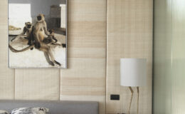 Modern bedroom interior featuring a gray bed with purple bedding, decorative pillows, a gold side table with a unique lamp, and a nature-themed artwork of monkeys hanging on textured beige walls.