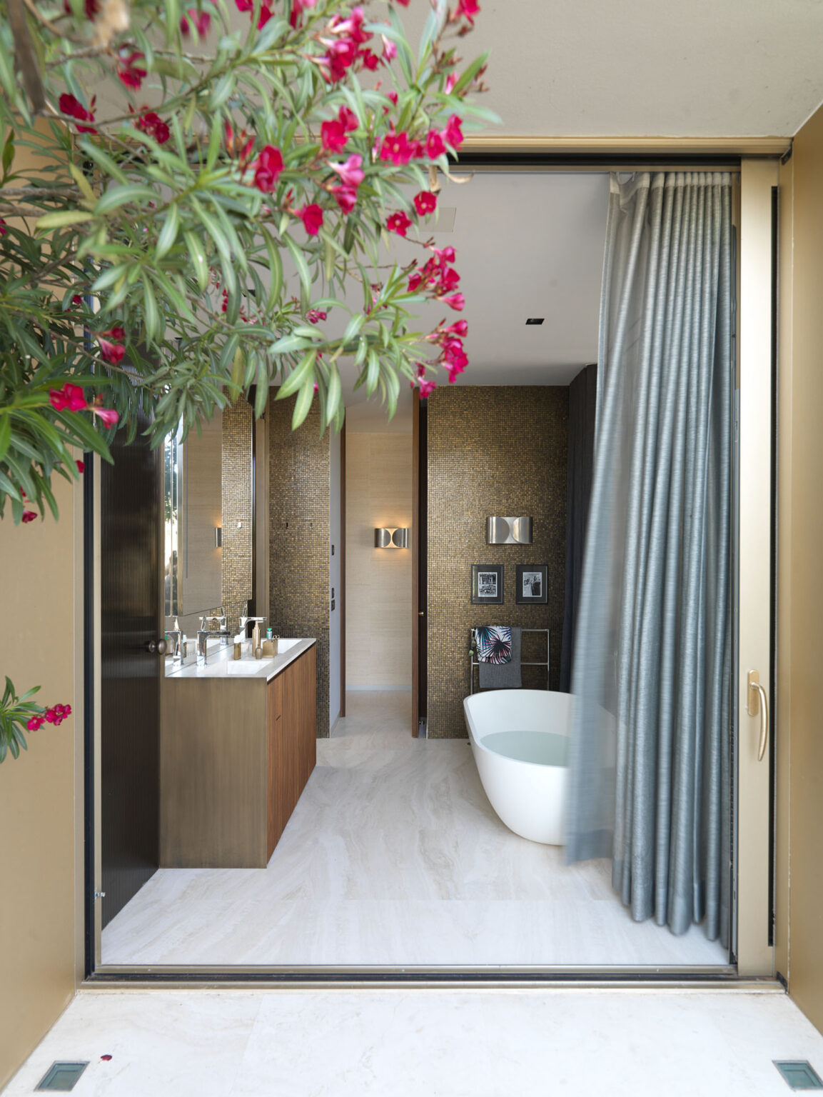Modern bathroom design featuring a freestanding bathtub, dual vanity with sleek fixtures, and elegant decor. The space is illuminated by warm lighting, with a view framed by lush greenery and vibrant pink flowers. | Only on SopranoVillas