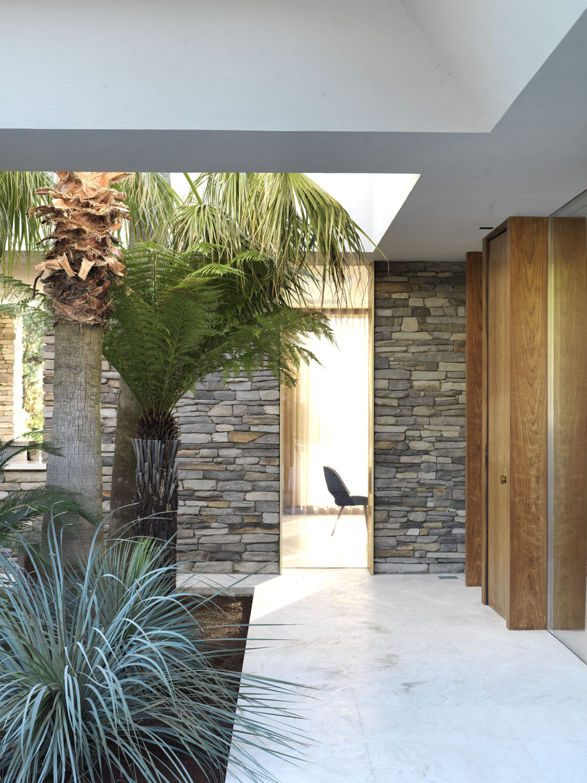 Interior view of a modern architectural space featuring a combination of stone walls, tropical plants, and a minimalist design. A glimpse of a chair is visible through a doorway, with natural light illuminating the area. | Only on SopranoVillas