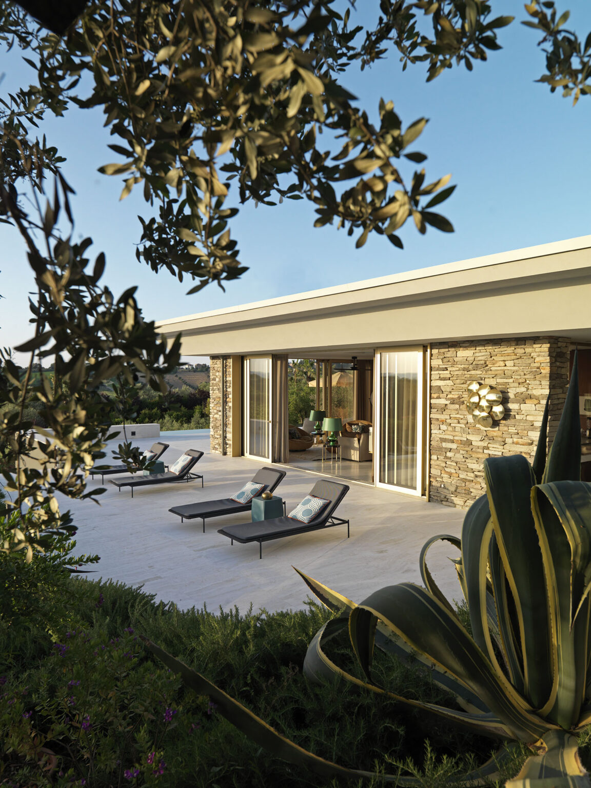 Modern villa exterior with a stone facade, featuring lounge chairs and a patio surrounded by lush greenery and plants, set against a clear blue sky. | Only on SopranoVillas