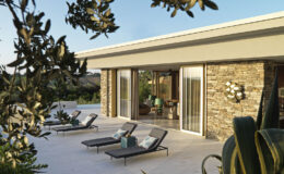 Modern villa exterior with a stone facade, featuring lounge chairs and a patio surrounded by lush greenery and plants, set against a clear blue sky.