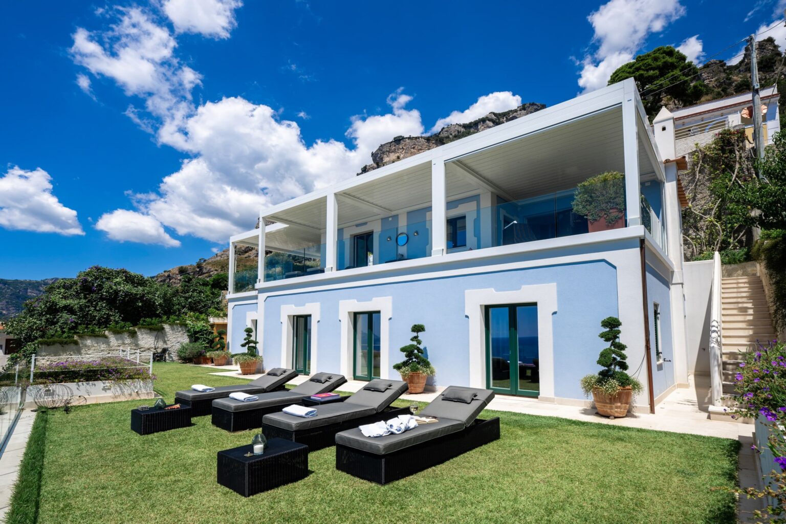 Modern luxury villa with a blue exterior, large glass windows, and a spacious terrace featuring sun loungers, set against a backdrop of lush greenery and a bright blue sky. | Only on SopranoVillas