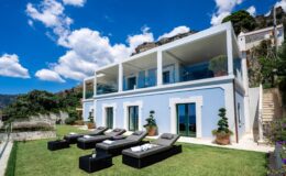 Modern luxury villa with a blue exterior, large glass windows, and a spacious terrace featuring sun loungers, set against a backdrop of lush greenery and a bright blue sky.