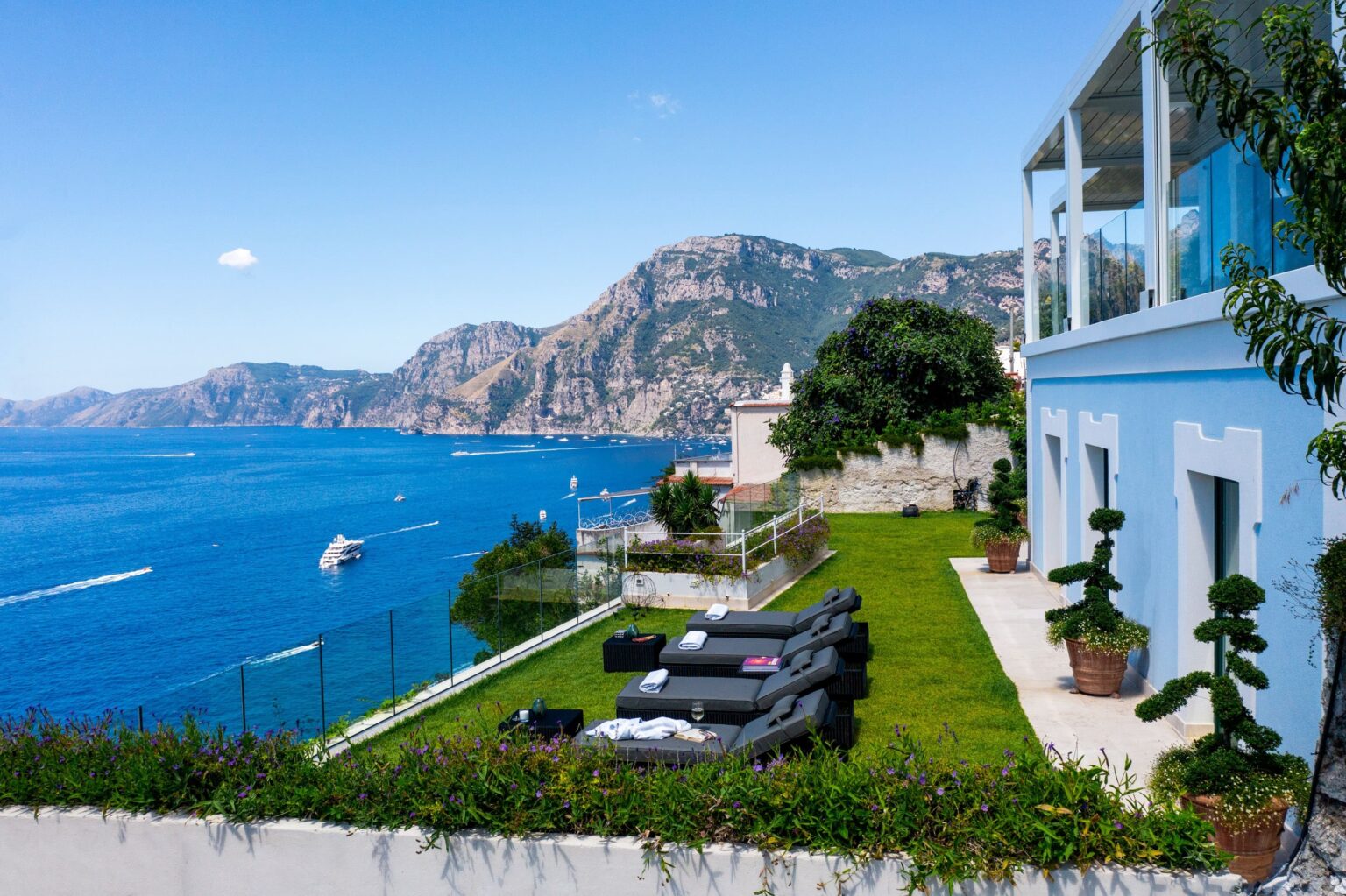 Luxurious seaside villa overlooking the ocean with stunning mountain views, featuring a green lawn and stylish lounge chairs for relaxation. | Only on SopranoVillas