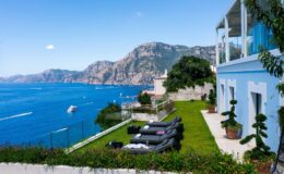 Luxurious seaside villa overlooking the ocean with stunning mountain views, featuring a green lawn and stylish lounge chairs for relaxation.