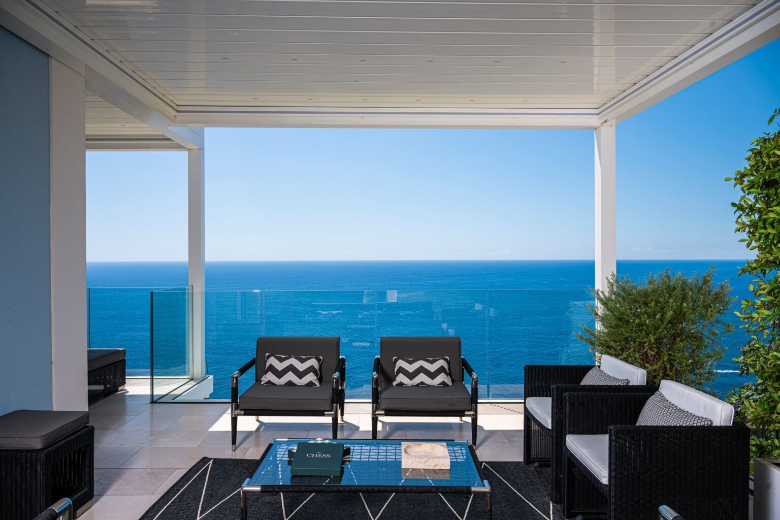 Modern outdoor living space with stylish black and white furniture, overlooking a stunning ocean view. The area is shaded by a sleek canopy, featuring a glass railing and potted greenery, creating a serene and inviting atmosphere. Perfect for relaxation and enjoying coastal scenery. | Only on SopranoVillas