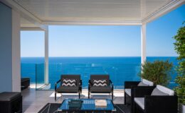 Modern outdoor living space with stylish black and white furniture, overlooking a stunning ocean view. The area is shaded by a sleek canopy, featuring a glass railing and potted greenery, creating a serene and inviting atmosphere. Perfect for relaxation and enjoying coastal scenery.