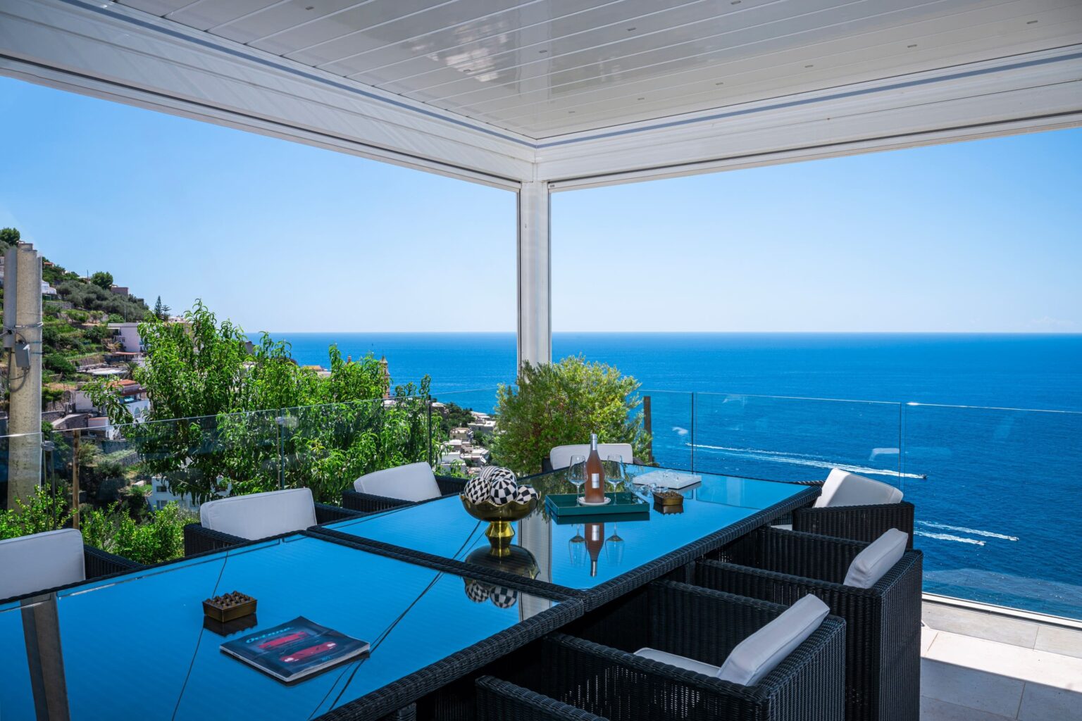 Modern outdoor dining area with a glass table and comfortable chairs, overlooking a stunning ocean view. Lush greenery frames the scene, creating a tranquil and inviting atmosphere for alfresco dining. | Only on SopranoVillas