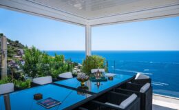 Modern outdoor dining area with a glass table and comfortable chairs, overlooking a stunning ocean view. Lush greenery frames the scene, creating a tranquil and inviting atmosphere for alfresco dining.