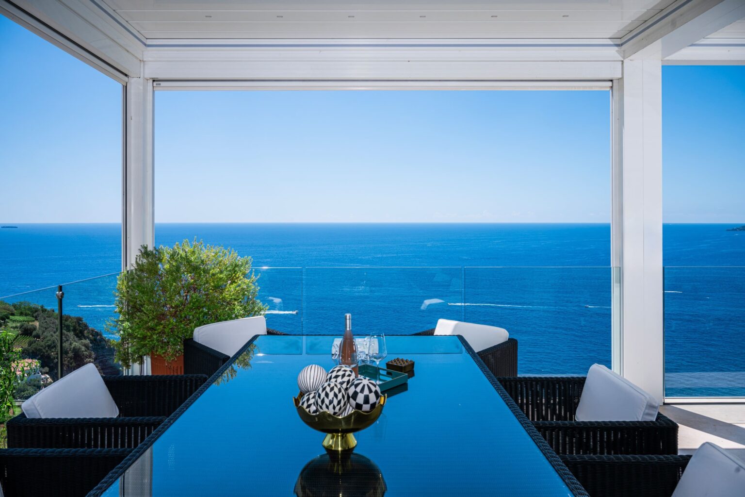 Modern dining area with a glass table overlooking a stunning ocean view, featuring decorative elements like a bowl and potted plant in a bright, airy environment. | Only on SopranoVillas