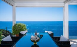 Modern dining area with a glass table overlooking a stunning ocean view, featuring decorative elements like a bowl and potted plant in a bright, airy environment.