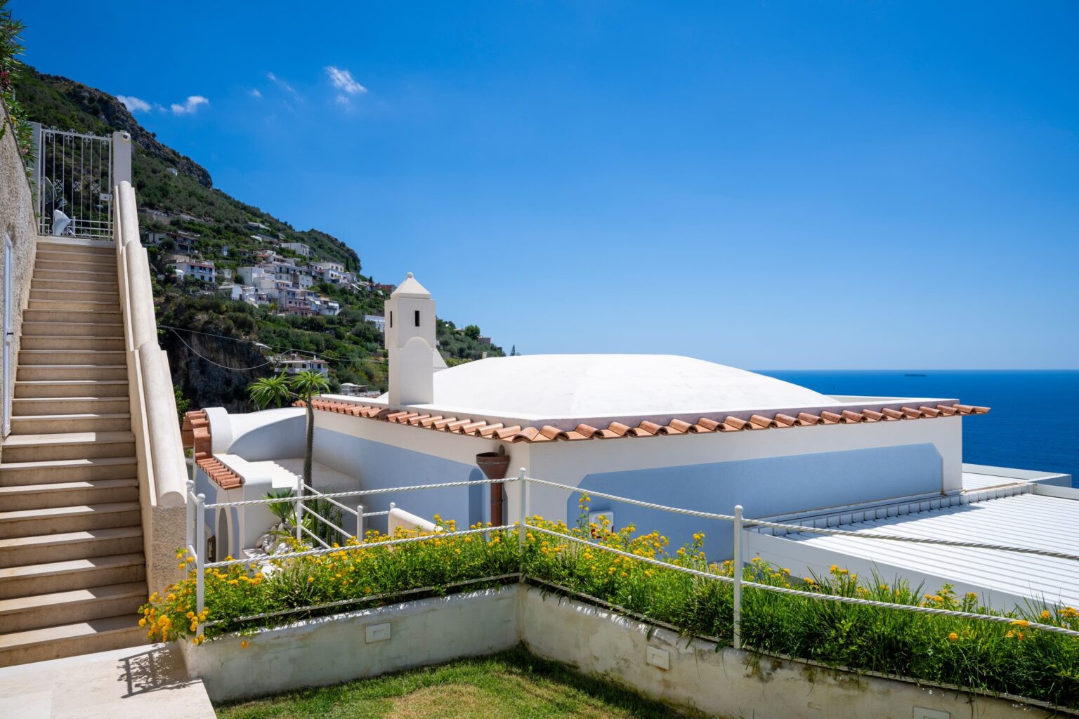 Modern villa with a white dome roof, surrounded by lush greenery and a staircase leading to a terrace overlooking the blue sea and sky, located in a coastal area. | Only on SopranoVillas
