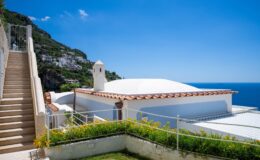 Modern villa with a white dome roof, surrounded by lush greenery and a staircase leading to a terrace overlooking the blue sea and sky, located in a coastal area.