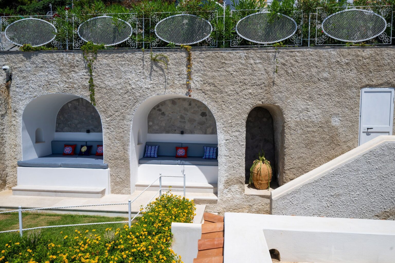 Cozy outdoor seating areas with white stone walls and colorful cushions, surrounded by lush greenery and decorative planters, ideal for relaxation in a Mediterranean-style garden. | Only on SopranoVillas