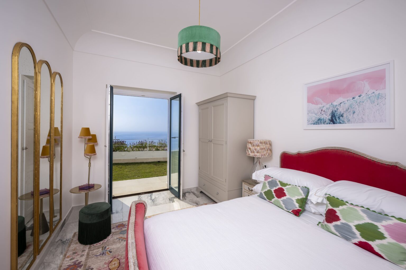 Cozy bedroom featuring a double bed with colorful patterned pillows, a stylish green pendant light, and a scenic view through an open door leading to a garden. The room includes a wardrobe, a decorative lamp, and a large mirror, creating a bright and inviting atmosphere. | Only on SopranoVillas
