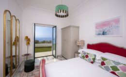 Cozy bedroom featuring a double bed with colorful patterned pillows, a stylish green pendant light, and a scenic view through an open door leading to a garden. The room includes a wardrobe, a decorative lamp, and a large mirror, creating a bright and inviting atmosphere.