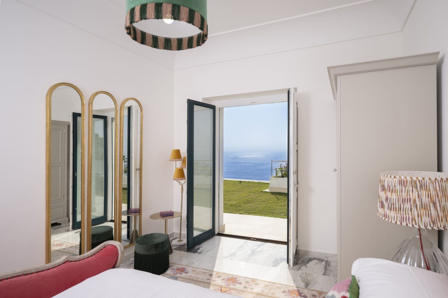 Modern bedroom interior with large open doors revealing a scenic view of the ocean. Features stylish decor including a pink bed, decorative mirrors, and contemporary lighting. Bright and airy atmosphere with natural light streaming in. | Only on SopranoVillas