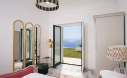 Modern bedroom interior with large open doors revealing a scenic view of the ocean. Features stylish decor including a pink bed, decorative mirrors, and contemporary lighting. Bright and airy atmosphere with natural light streaming in.