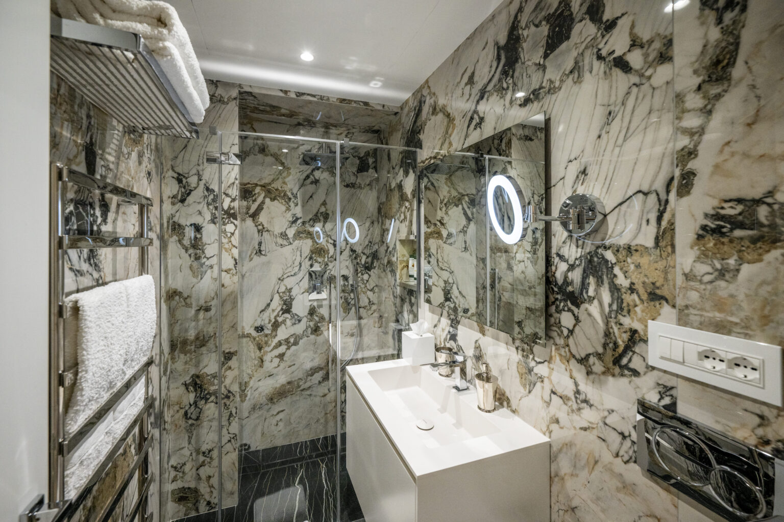 Modern bathroom featuring elegant marble walls, a glass shower enclosure, a sleek white vanity with contemporary fixtures, and illuminated mirrors, creating a luxurious and spacious atmosphere. | Only on SopranoVillas