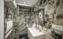 Modern bathroom featuring elegant marble walls, a glass shower enclosure, a sleek white vanity with contemporary fixtures, and illuminated mirrors, creating a luxurious and spacious atmosphere.
