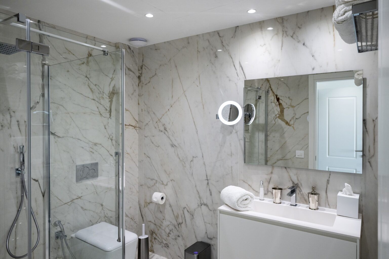 Modern bathroom interior featuring elegant marble walls, a glass shower enclosure, a sleek vanity with toiletries, and bright lighting for a stylish and functional design. | Only on SopranoVillas