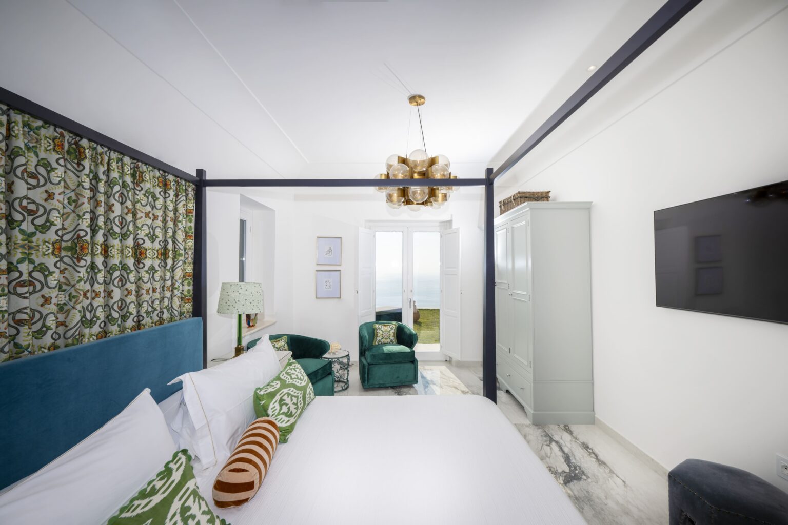 Modern hotel room featuring a stylish four-poster bed, decorative pillows, green accent chairs, and contemporary decor, with large windows allowing natural light and a television mounted on the wall. | Only on SopranoVillas