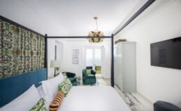 Modern hotel room featuring a stylish four-poster bed, decorative pillows, green accent chairs, and contemporary decor, with large windows allowing natural light and a television mounted on the wall.