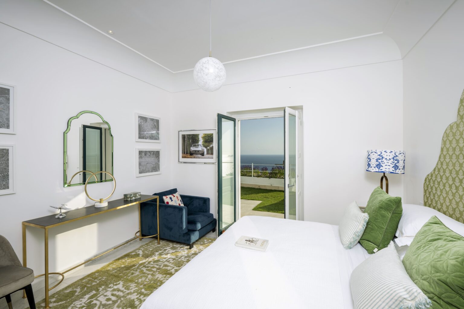 Bright and airy bedroom featuring a king-size bed with green decorative pillows, a cozy blue armchair, and a stylish vanity. Large glass doors open to a scenic view of the ocean, enhancing the room's serene atmosphere. | Only on SopranoVillas