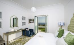Bright and airy bedroom featuring a king-size bed with green decorative pillows, a cozy blue armchair, and a stylish vanity. Large glass doors open to a scenic view of the ocean, enhancing the room's serene atmosphere.