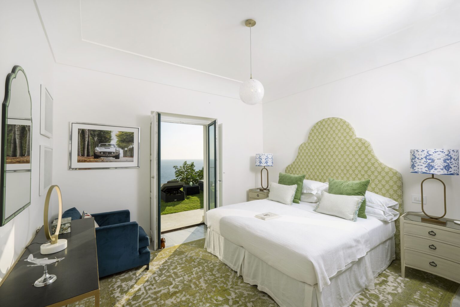Modern hotel bedroom with a large double bed, green accent pillows, and stylish decor. Features include a cozy seating area, large windows with a scenic view, and elegant wall mirrors, creating a bright and inviting atmosphere. | Only on SopranoVillas