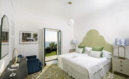 Modern hotel bedroom with a large double bed, green accent pillows, and stylish decor. Features include a cozy seating area, large windows with a scenic view, and elegant wall mirrors, creating a bright and inviting atmosphere.