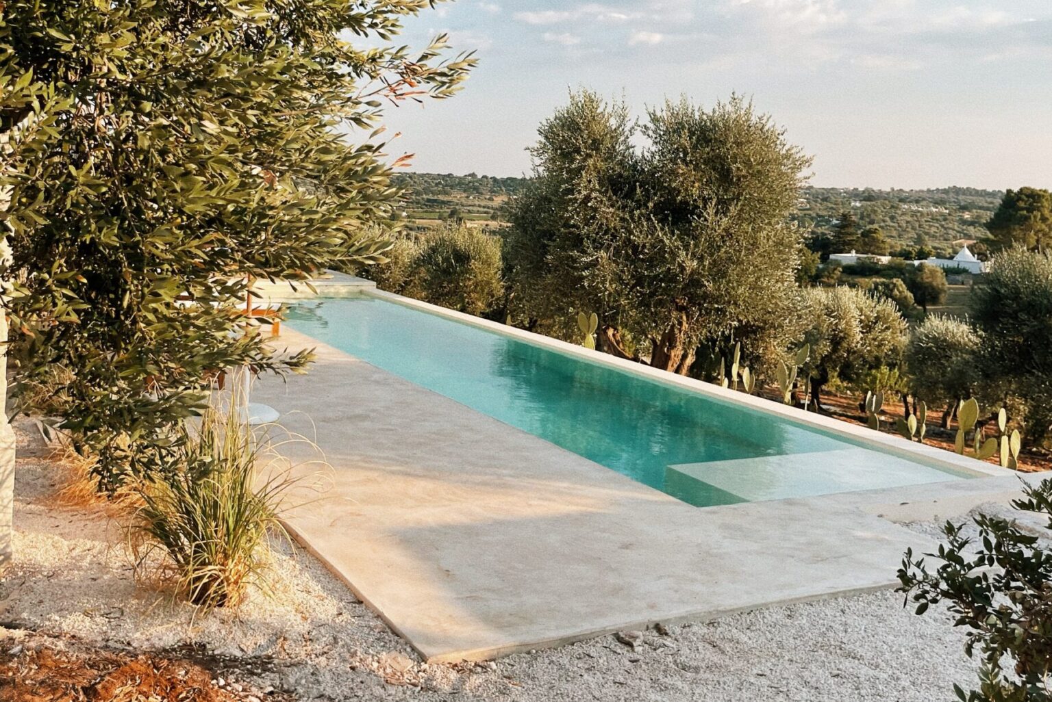 Modern infinity pool surrounded by olive trees and lush greenery, set against a scenic hillside landscape. The serene outdoor space features a sleek concrete deck and tranquil water, ideal for relaxation and leisure. | Only on SopranoVillas