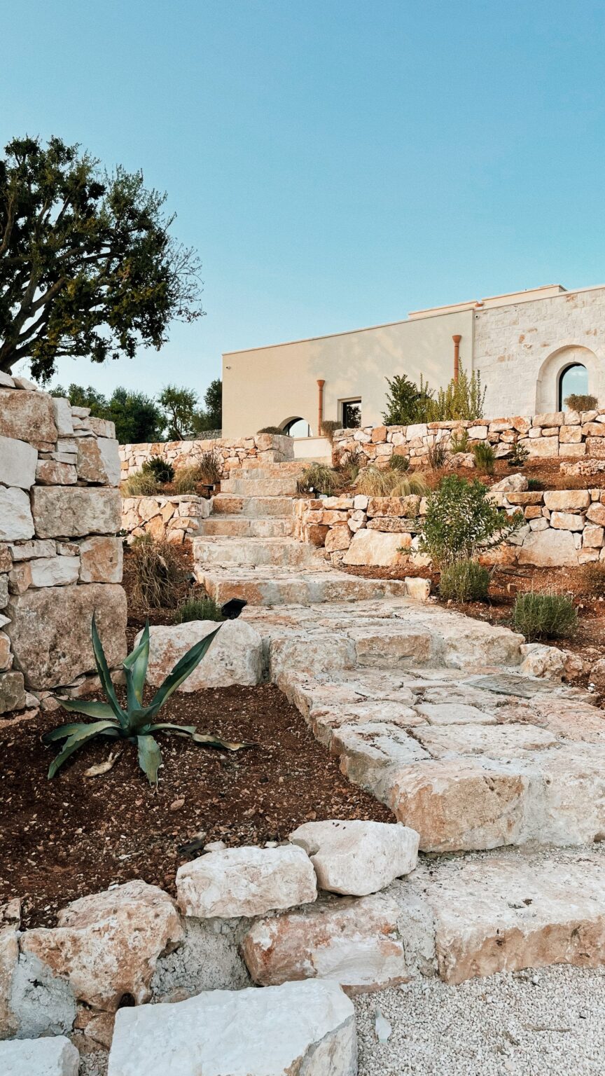Stone pathway leading up through a landscaped garden with native plants and a modern building in the background, under a clear blue sky. | Only on SopranoVillas