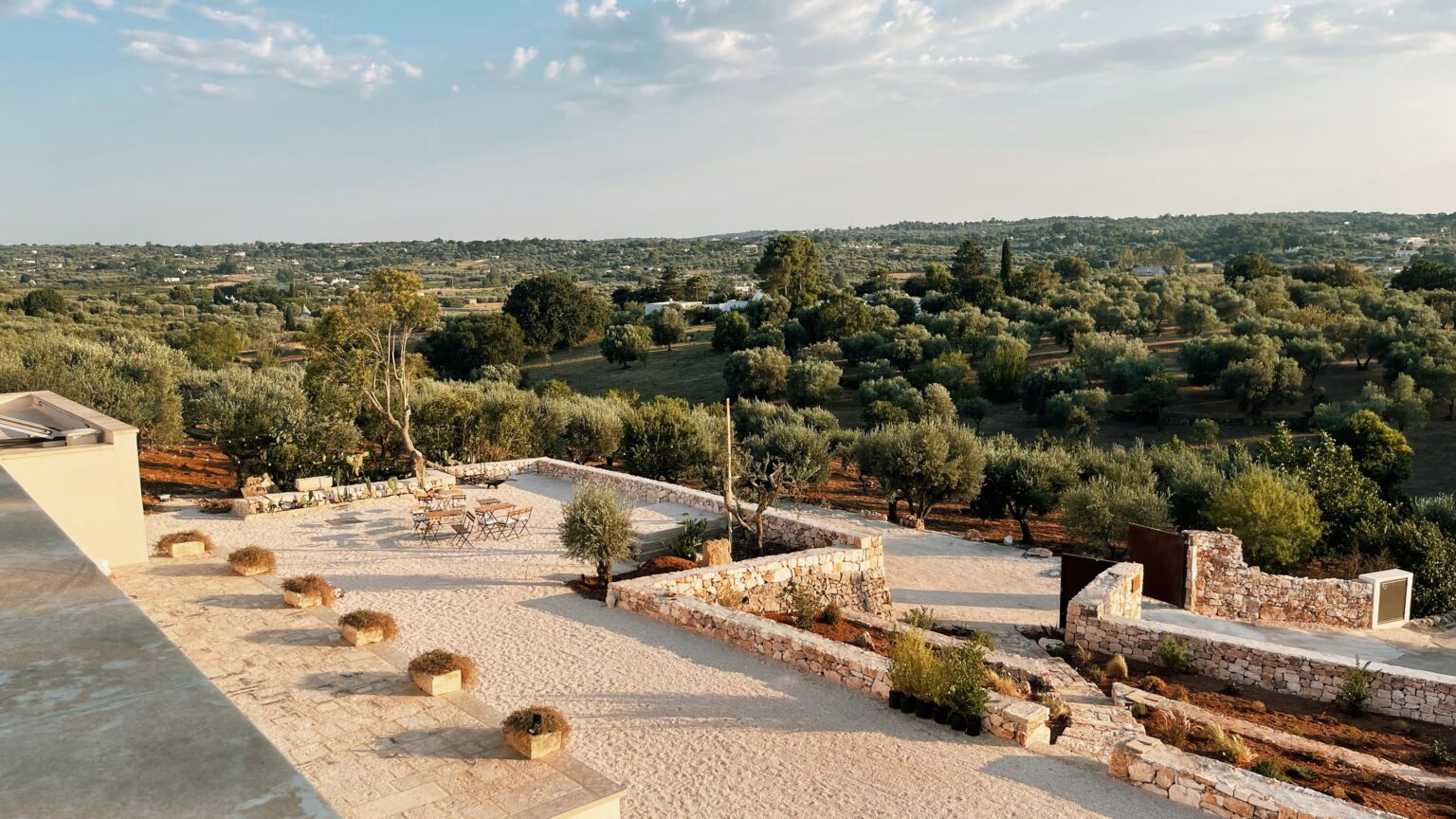 Scenic view of a landscaped outdoor area featuring stone pathways and seating arrangements, surrounded by lush green olive trees and rolling hills under a clear blue sky. Perfect for relaxation and enjoying nature. | Only on SopranoVillas