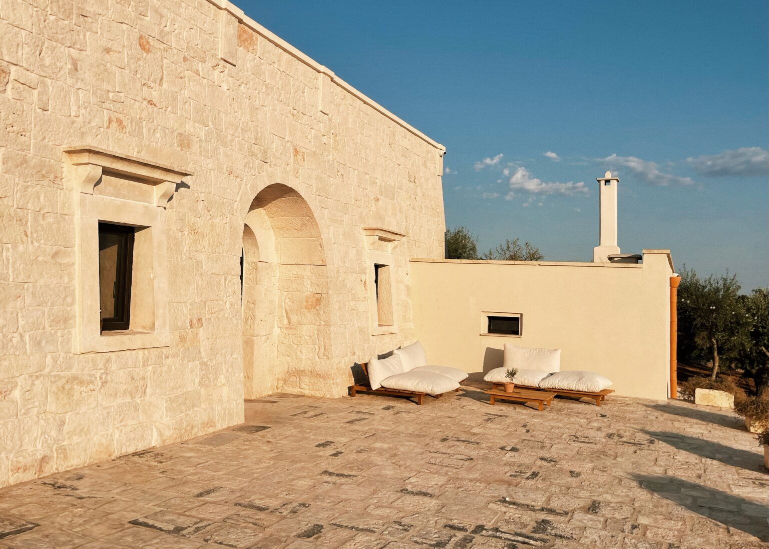 Exterior view of a rustic stone house featuring a large archway and two comfortable seating areas on a sunlit patio, surrounded by olive trees and a clear blue sky. | Only on SopranoVillas