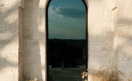 Arched doorway with a reflective glass panel surrounded by rustic stone walls, featuring potted plants and dried grasses at the base, set against a scenic landscape under a cloudy sky.