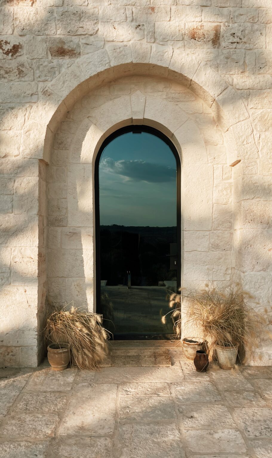 Arched doorway with a reflective glass panel surrounded by rustic stone walls, featuring potted plants and dried grasses at the base, set against a scenic landscape under a cloudy sky. | Only on SopranoVillas