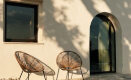 Modern exterior of a house featuring two stylish wicker chairs on a stone patio, with a large arched doorway and a window casting soft shadows from nearby trees.