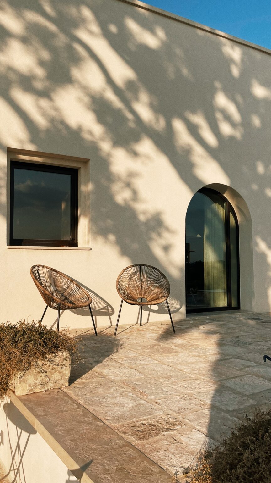 Modern exterior of a house featuring two stylish wicker chairs on a stone patio, with a large arched doorway and a window casting soft shadows from nearby trees. | Only on SopranoVillas