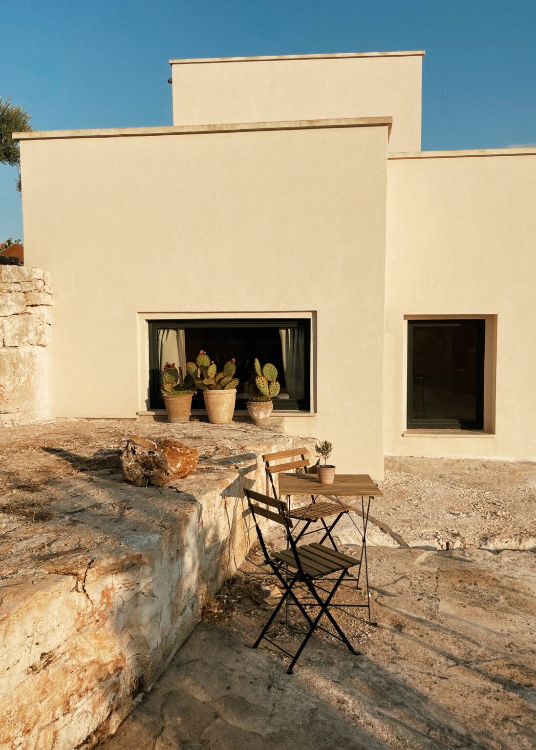 Modern minimalist house exterior with a stone terrace, featuring potted cacti and a small wooden table with chairs, under a clear blue sky. | Only on SopranoVillas
